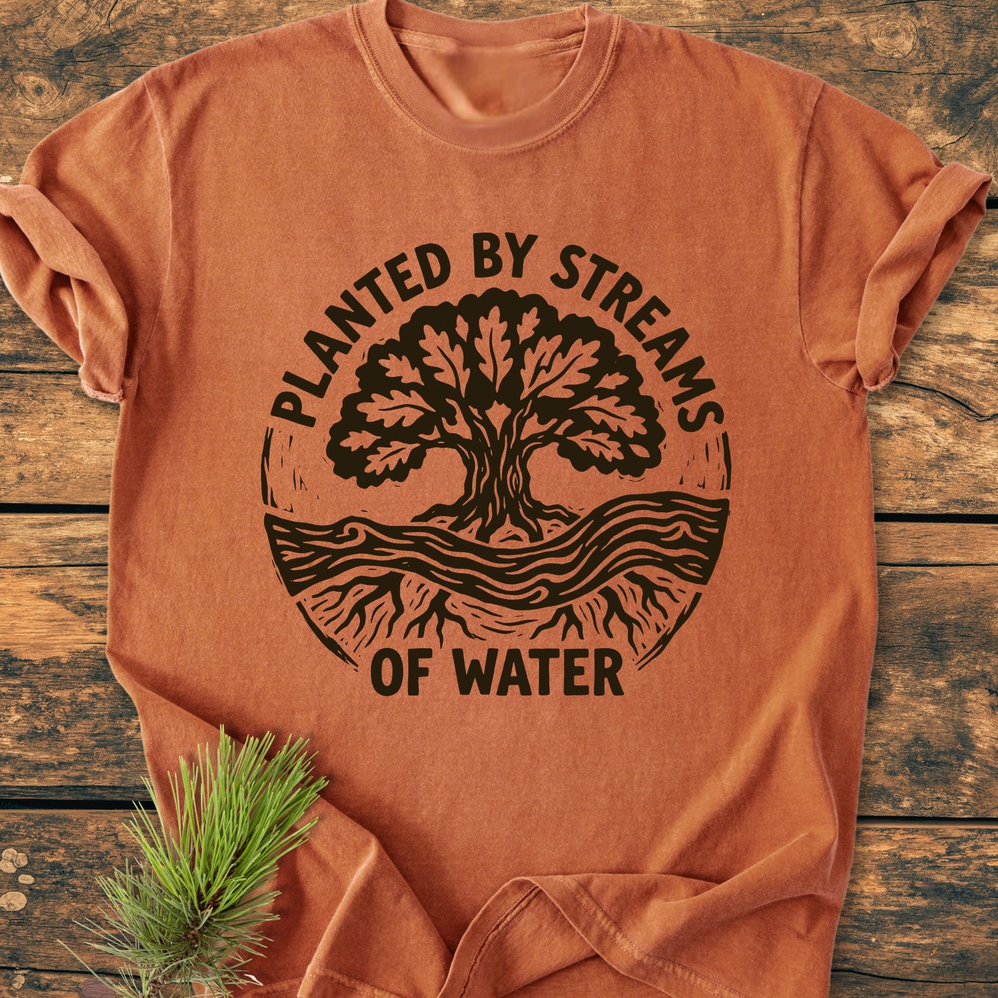 Planted By Streams, Psalm 1,3 - Vintage Wash Tee features a woodcut-style tree design with roots and stream on a burnt-orange T-shirt, laid on rustic wood with a pine sprig beside it.