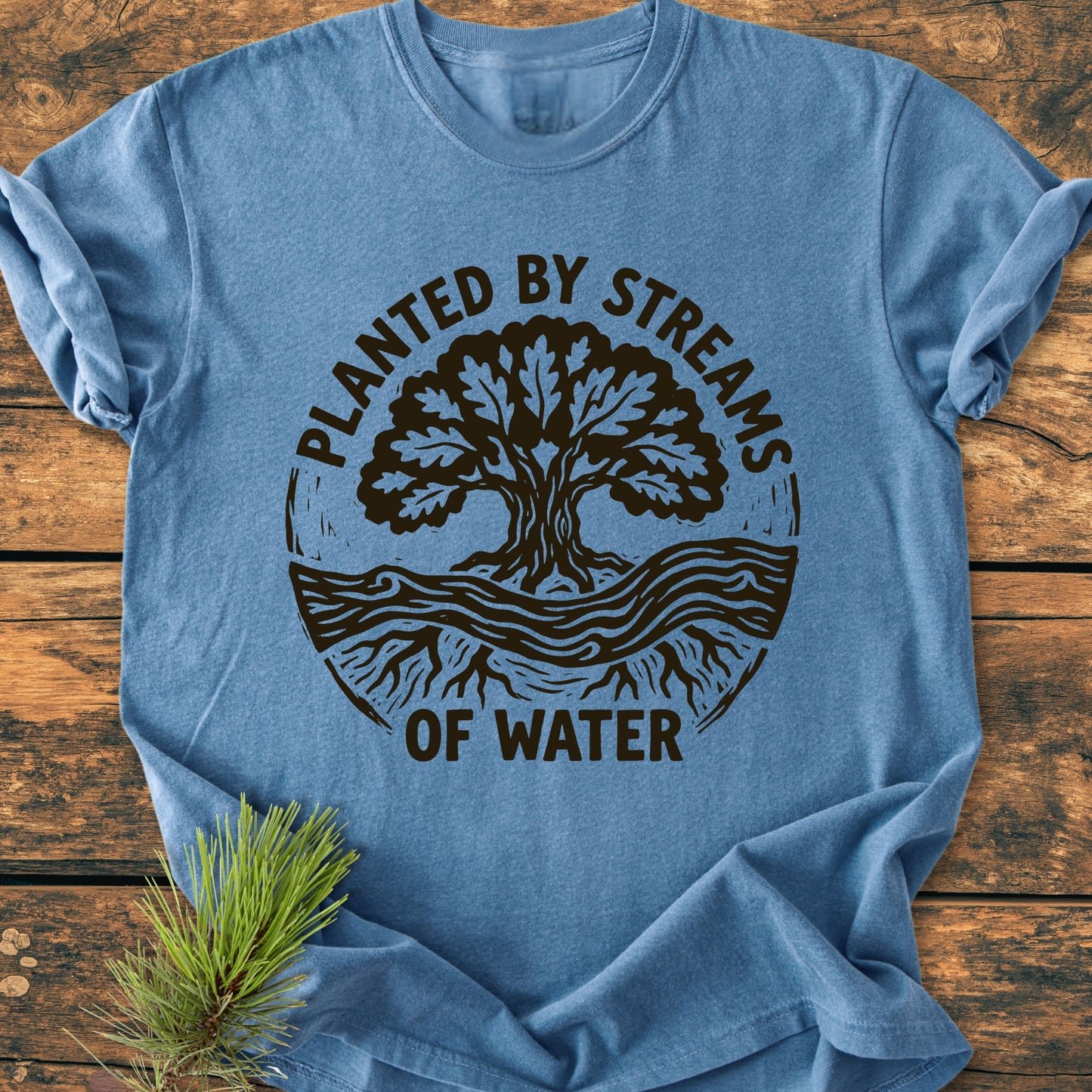 Heather-blue Planted By Streams, Psalm 1,3 - Vintage Wash Tee with tree emblem and text, laid on wood. Features soft-washed, garment-dyed fabric for comfort and durability. Pine sprig accents image.