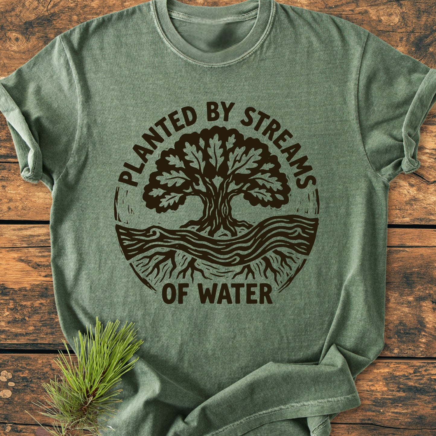 Heather-green Planted By Streams, Psalm 1,3 - Vintage Wash Tee featuring a circular tree and water graphic, laid on wood with a pine sprig, showcasing a relaxed fit and durable design.