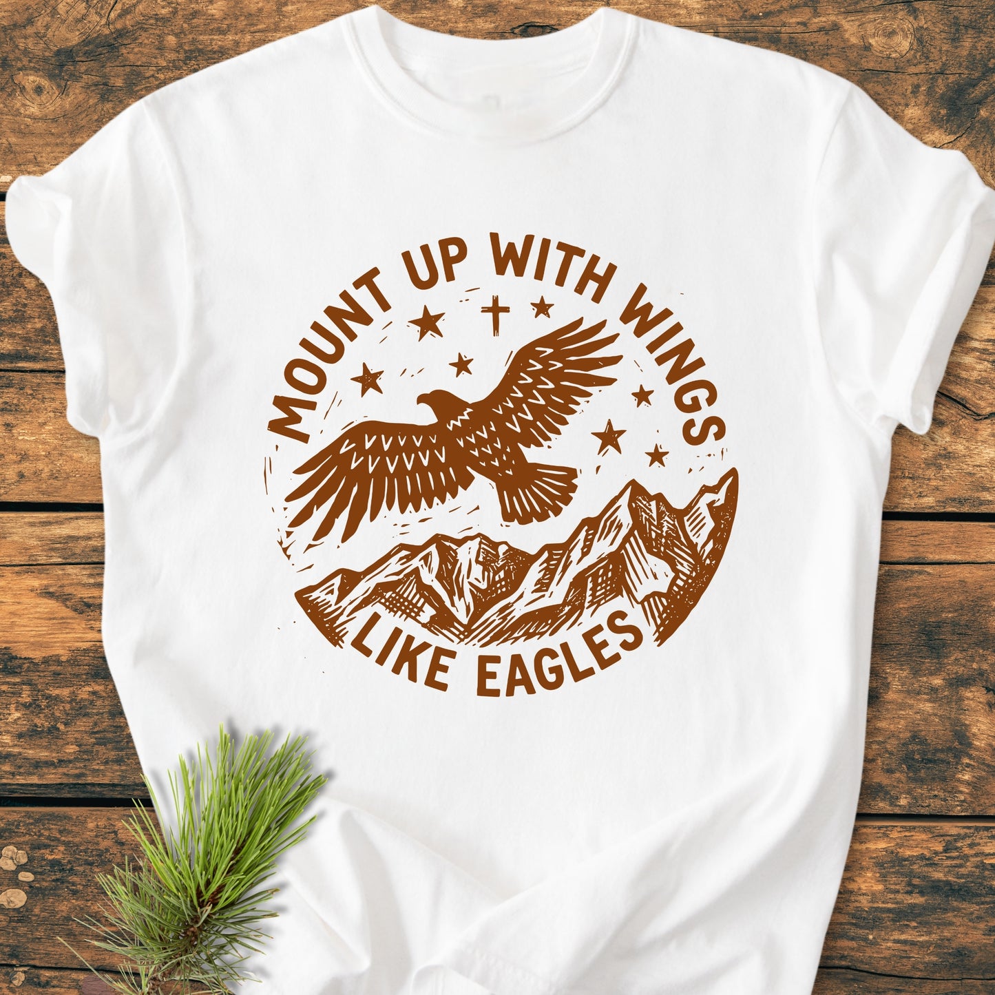 Mount Up With Wings - Vintage Wash Tee featuring a brown-ink design of an eagle, mountain range, and stars, with the slogan, laid on a wooden surface. Made from soft, pre-shrunk ring-spun cotton.