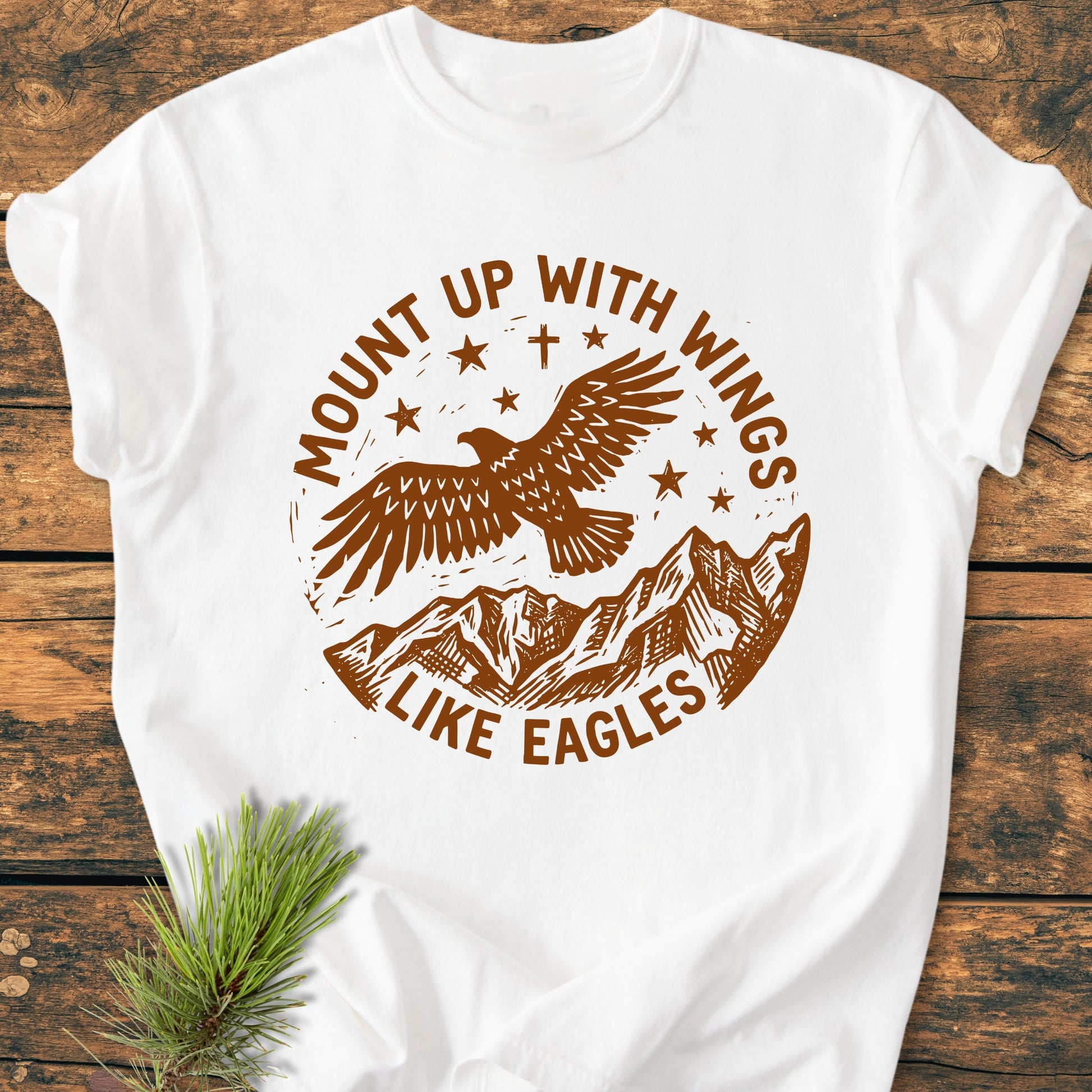 Mount Up With Wings - Vintage Wash Tee featuring a brown-ink design of an eagle, mountain range, and stars, with the slogan, laid on a wooden surface. Made from soft, pre-shrunk ring-spun cotton.