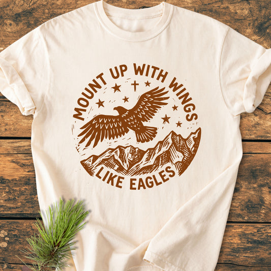 Mount Up With Wings - Vintage Wash Tee featuring an eagle graphic and stars, laid flat on wood. Cream-colored, 100% cotton, relaxed fit. Includes pine sprig accent.