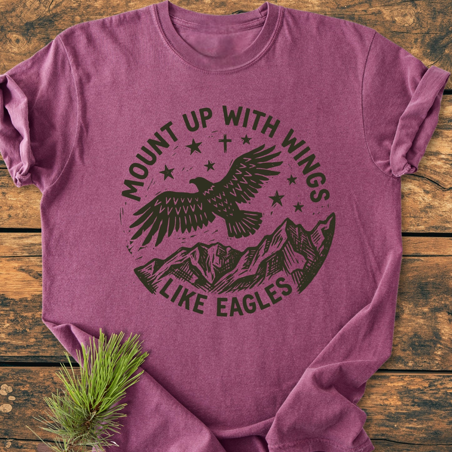 Mount Up With Wings - Vintage Wash Tee features a heather-purple hue with a stamp-style graphic of an eagle and mountains. Laid on wood, it includes a pine sprig detail for added rustic charm.