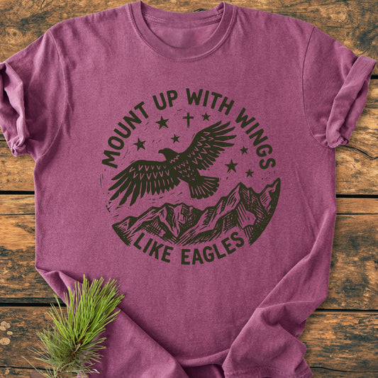 Mount Up With Wings - Vintage Wash Tee features a heather-purple hue with a stamp-style graphic of an eagle and mountains. Laid on wood, it includes a pine sprig detail for added rustic charm.