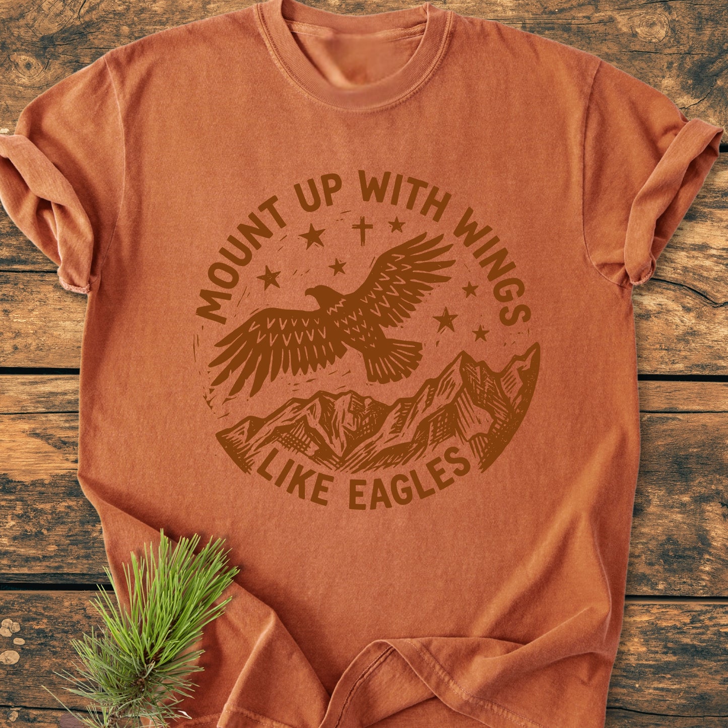 Mount Up With Wings - Vintage Wash Tee features a soaring eagle and mountain design, screen-printed on a rust-orange shirt, laid flat on wood, with a pine sprig accenting the display.