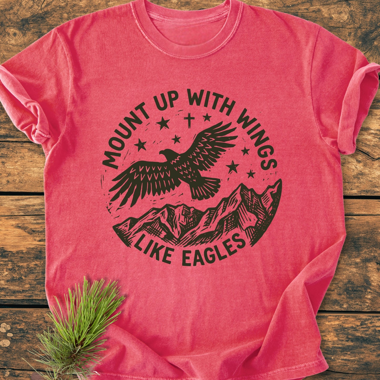 Mount Up With Wings - Vintage Wash Tee: A red, short-sleeve T-shirt featuring a black ink design of an eagle over mountains with stars, slogan arcing around, laid flat on a wooden surface.