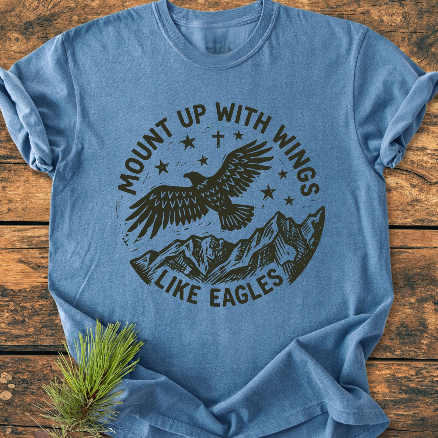 Mount Up With Wings - Vintage Wash Tee featuring a black eagle graphic, mountain range, and stars, laid flat. Heather-blue, 100% ring-spun cotton, relaxed fit, styled with pine sprig.