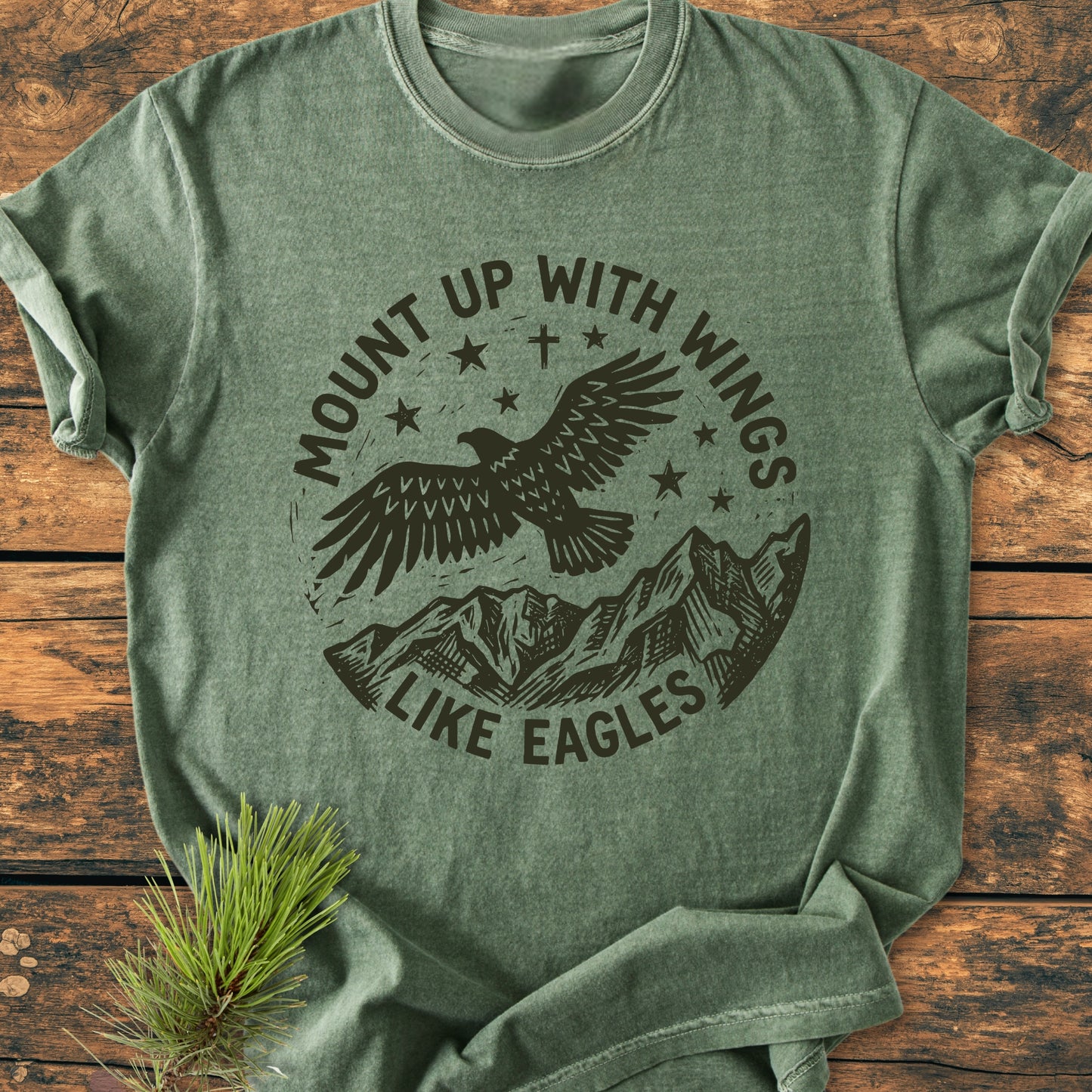 Mount Up With Wings - Vintage Wash Tee features a soaring eagle graphic with stars and a mountain range, screen-printed on a soft heather-green T-shirt, laid on a wooden surface with a pine sprig.