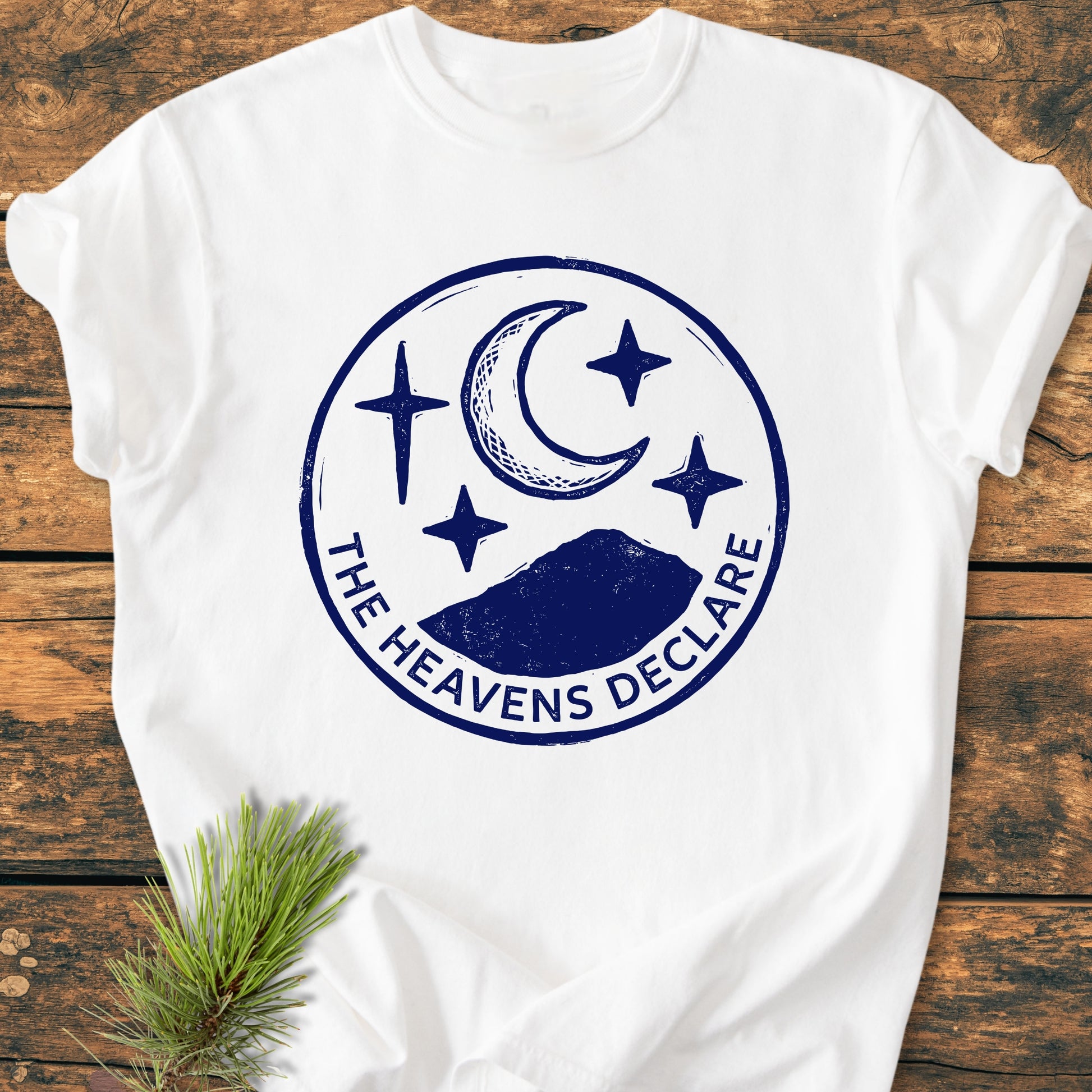 The Heavens Declare - Vintage Wash Tee featuring a navy-blue graphic of a mountain, crescent moon, and stars on soft, garment-dyed fabric, laid on rustic wood with pine decor.