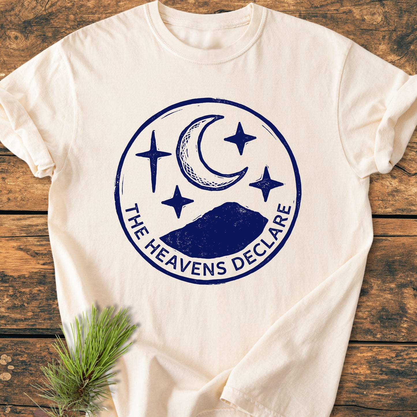 The Heavens Declare - Vintage Wash Tee features a navy circular graphic with a crescent moon, stars, and mountains on a cream shirt, evoking a natural, outdoorsy feel with an adjacent pine twig.