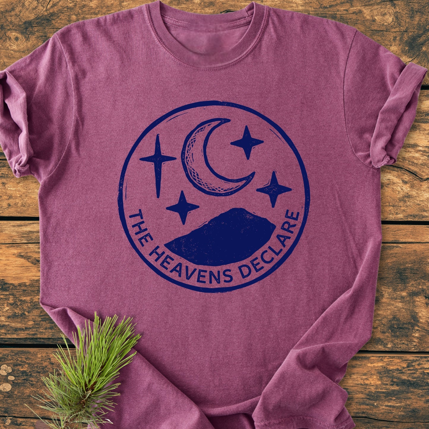 The Heavens Declare - Vintage Wash Tee: A purple-mauve T-shirt with a navy emblem featuring mountains, crescent moon, stars, and text. Laid on wood, a pine bough accents the scene.