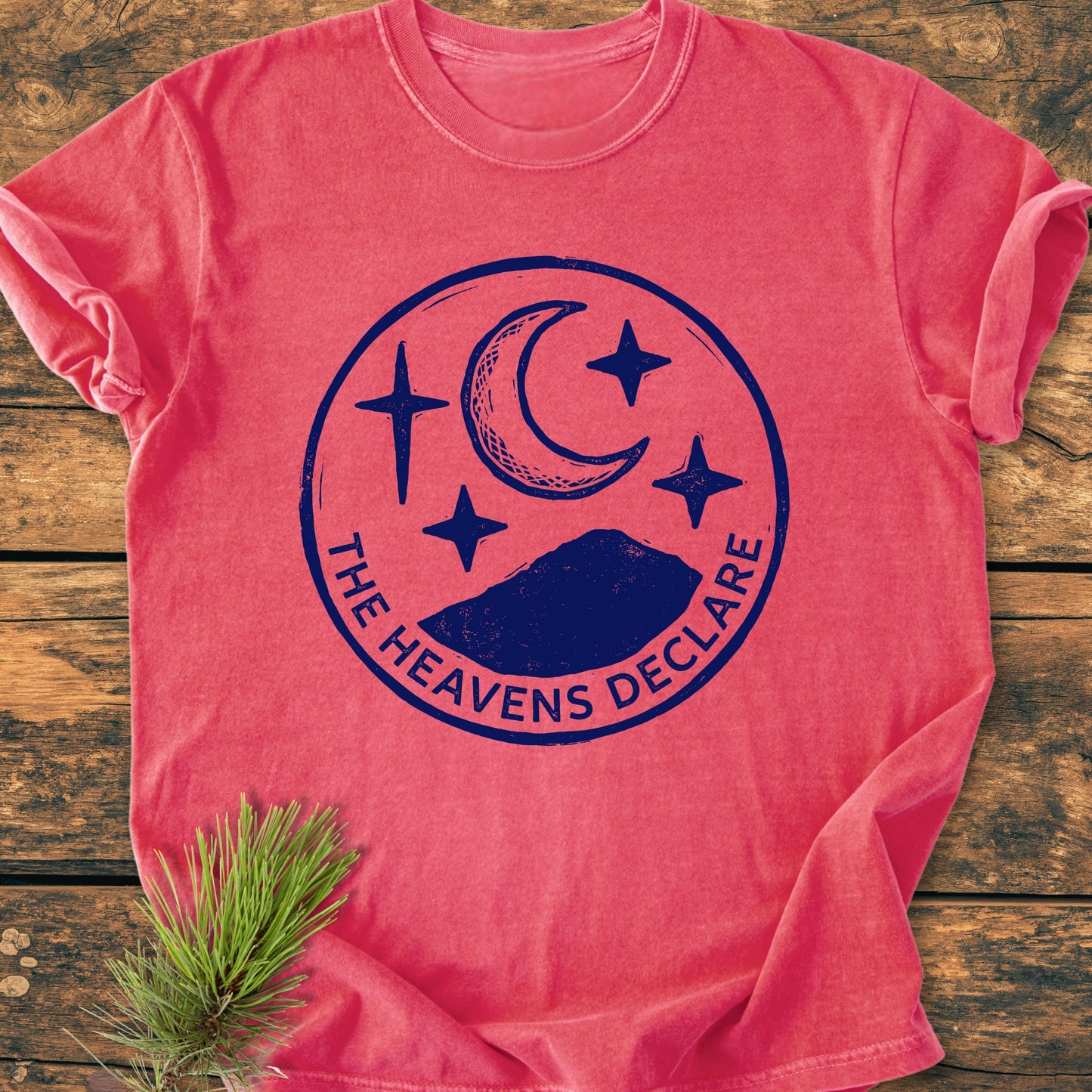 The Heavens Declare - Vintage Wash Tee, a heathered red, relaxed-fit shirt with a dark blue celestial emblem, laid flat on rustic wood, accented with a pine needle sprig.