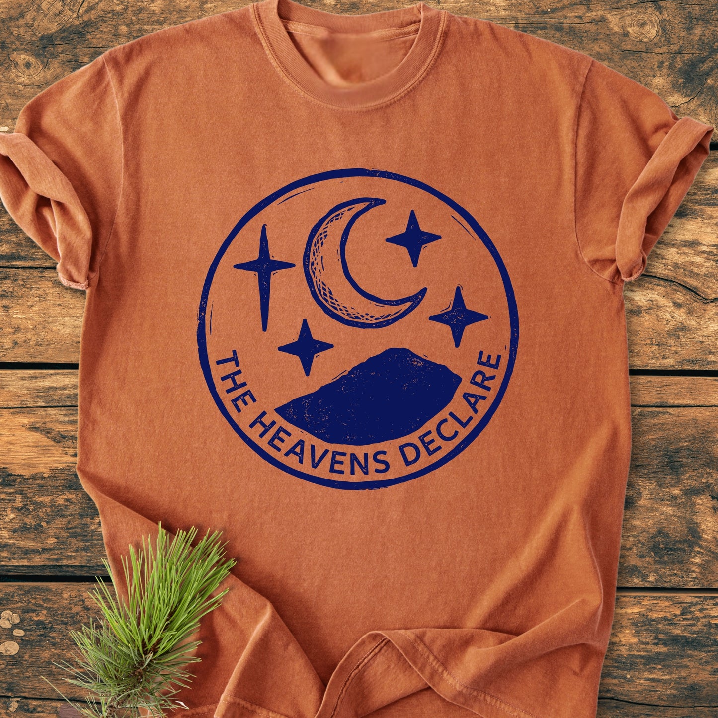 The Heavens Declare - Vintage Wash Tee displayed on wood, features a navy graphic of a crescent moon, stars, and mountains, with the phrase “THE HEAVENS DECLARE” beneath the circle design.