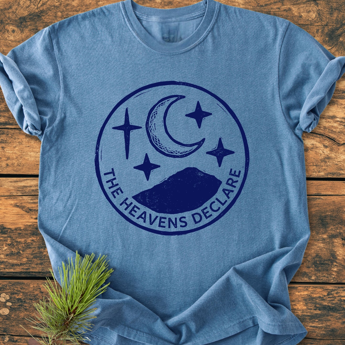 The Heavens Declare - Vintage Wash Tee features a heather-blue color with a stamp-style graphic of a crescent moon, stars, and mountains, laid flat on a wooden surface.