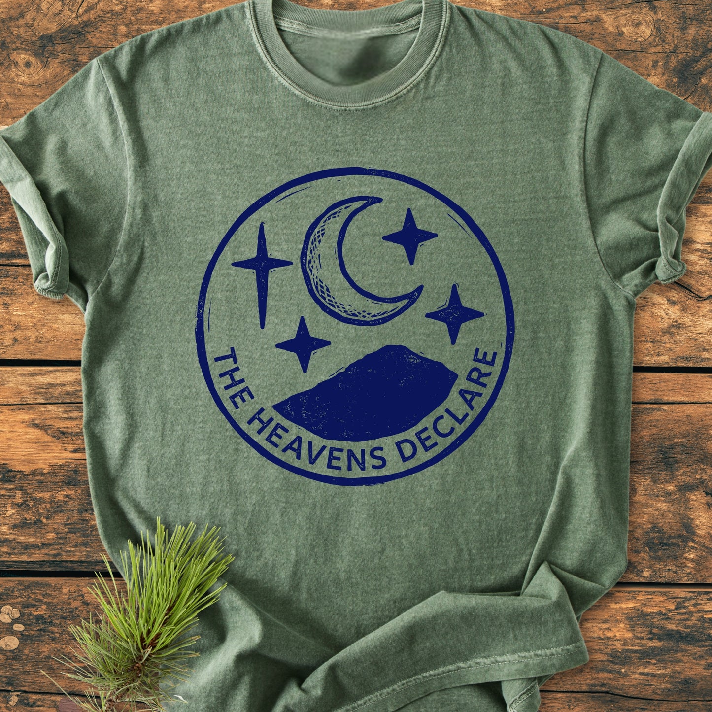 The Heavens Declare - Vintage Wash Tee features a soft green, short-sleeve style with a navy graphic of a crescent moon and stars, laid on rustic wood with a pine sprig accent.