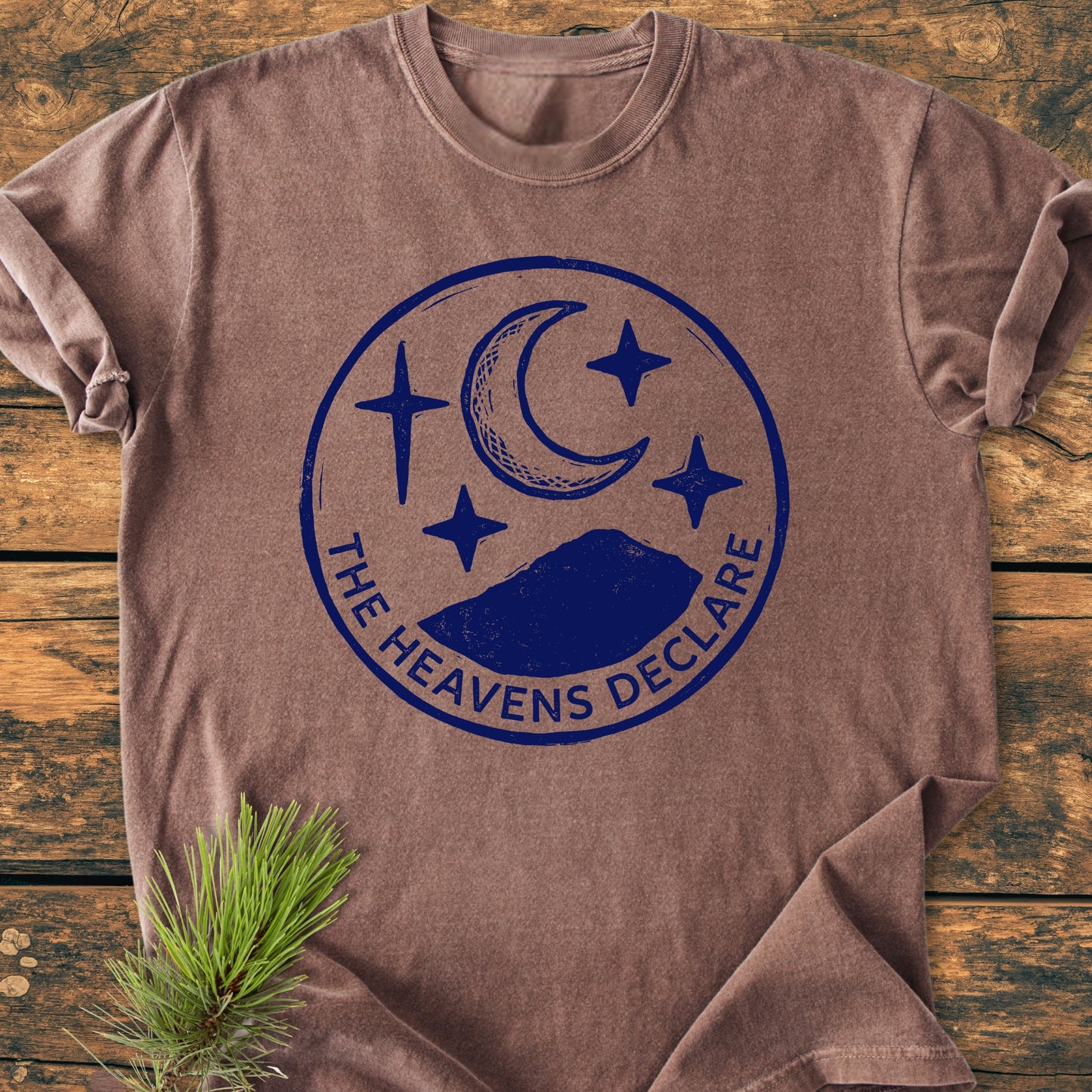 The Heavens Declare - Vintage Wash Tee features a navy circular graphic with a crescent moon, stars, and silhouetted hill, laid flat with a pine sprig accent for a rustic touch.
