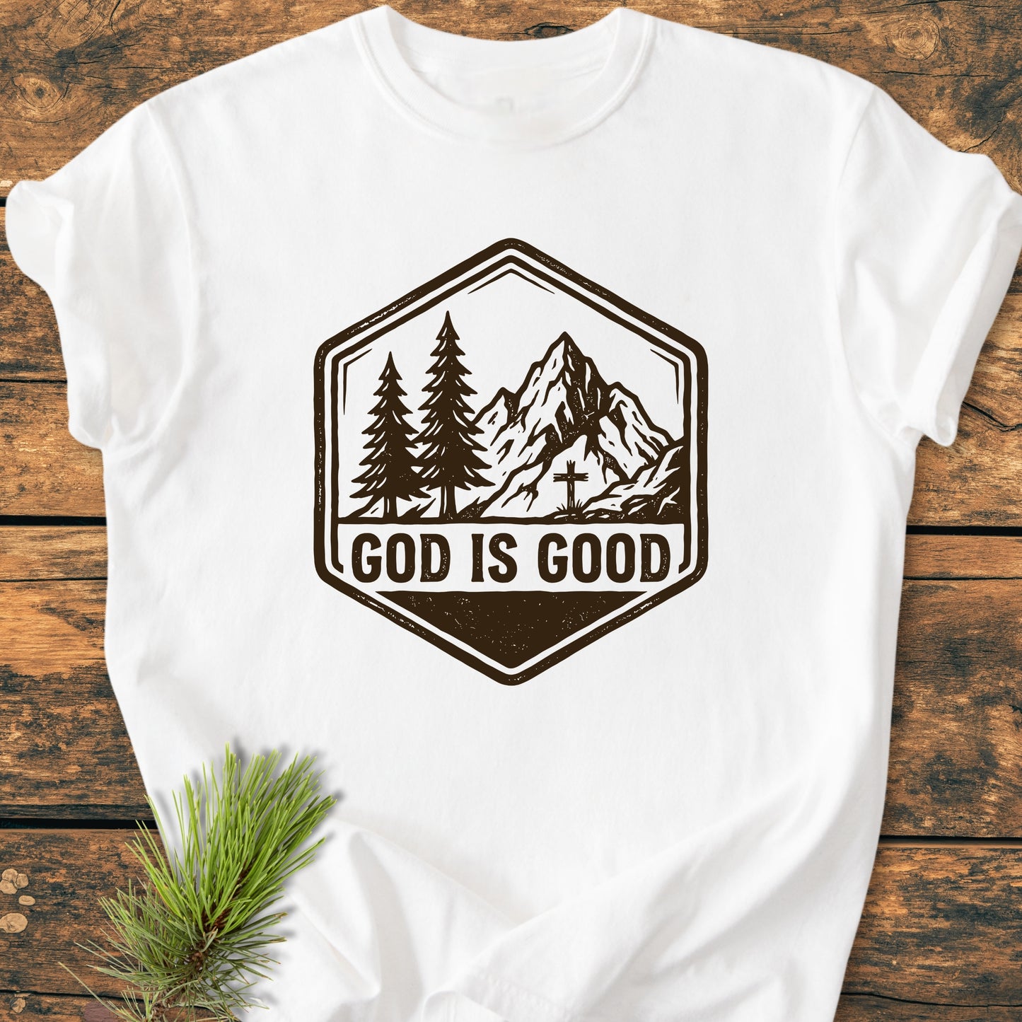 God Is Good - Vintage Wash Tee features a white shirt with a mountain and pine tree graphic, and bold GOD IS GOOD text, laid out on a wooden surface.