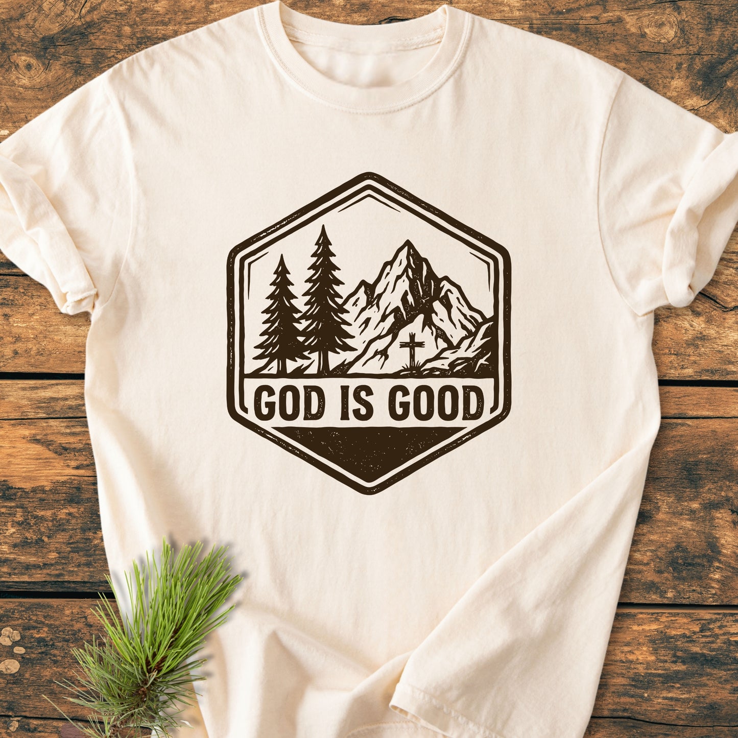 God Is Good - Vintage Wash Tee featuring a hexagonal badge with pine trees, a mountain peak, and a cross, styled with a pine sprig on a wooden surface.