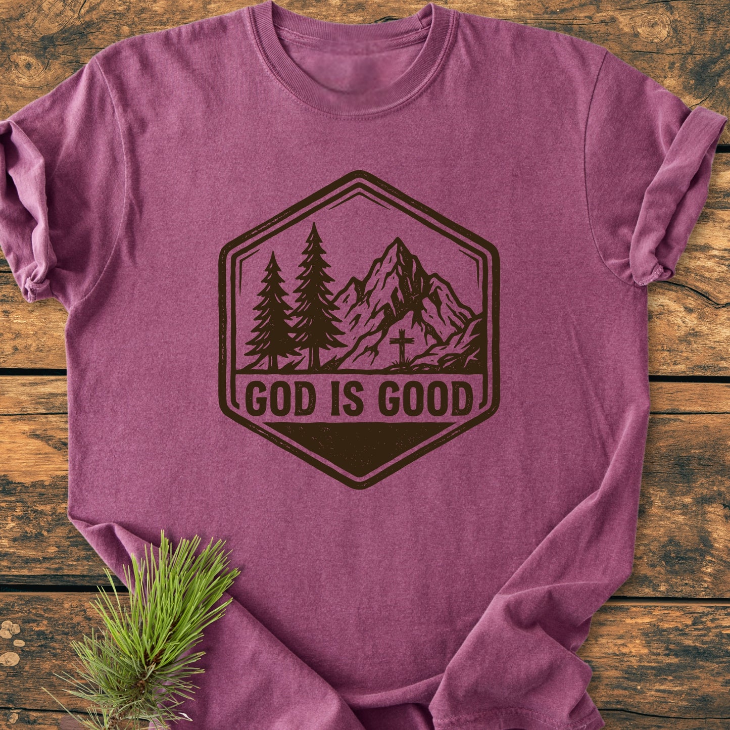 God Is Good - Vintage Wash Tee, featuring a badge-style graphic with pine trees and mountains, laid flat on wood. Includes a pine sprig accent, showcasing its relaxed fit and durable design.