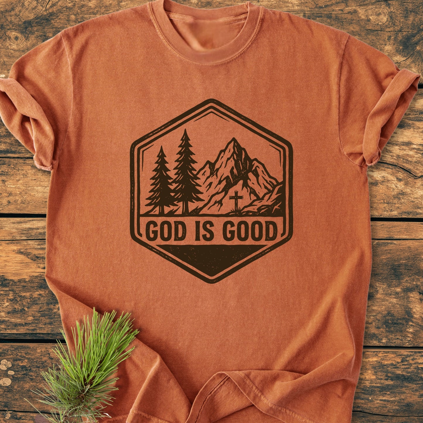 God Is Good - Vintage Wash Tee featuring a black mountain graphic and text, displayed on a wooden surface with pine needles, highlighting its relaxed fit and soft, garment-dyed cotton fabric.