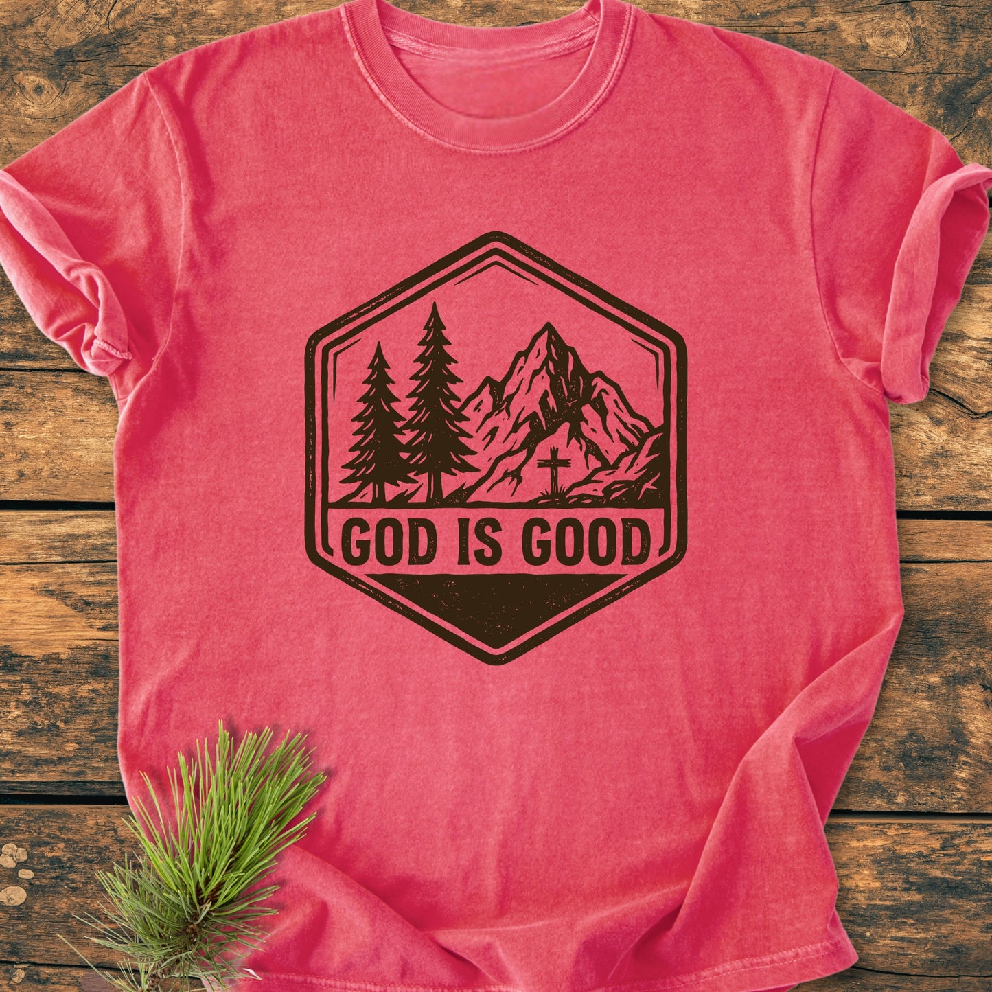 The God Is Good - Vintage Wash Tee features a badge graphic with pine trees and a mountain, laid flat on wood. It's a soft, relaxed fit, 100% cotton tee.