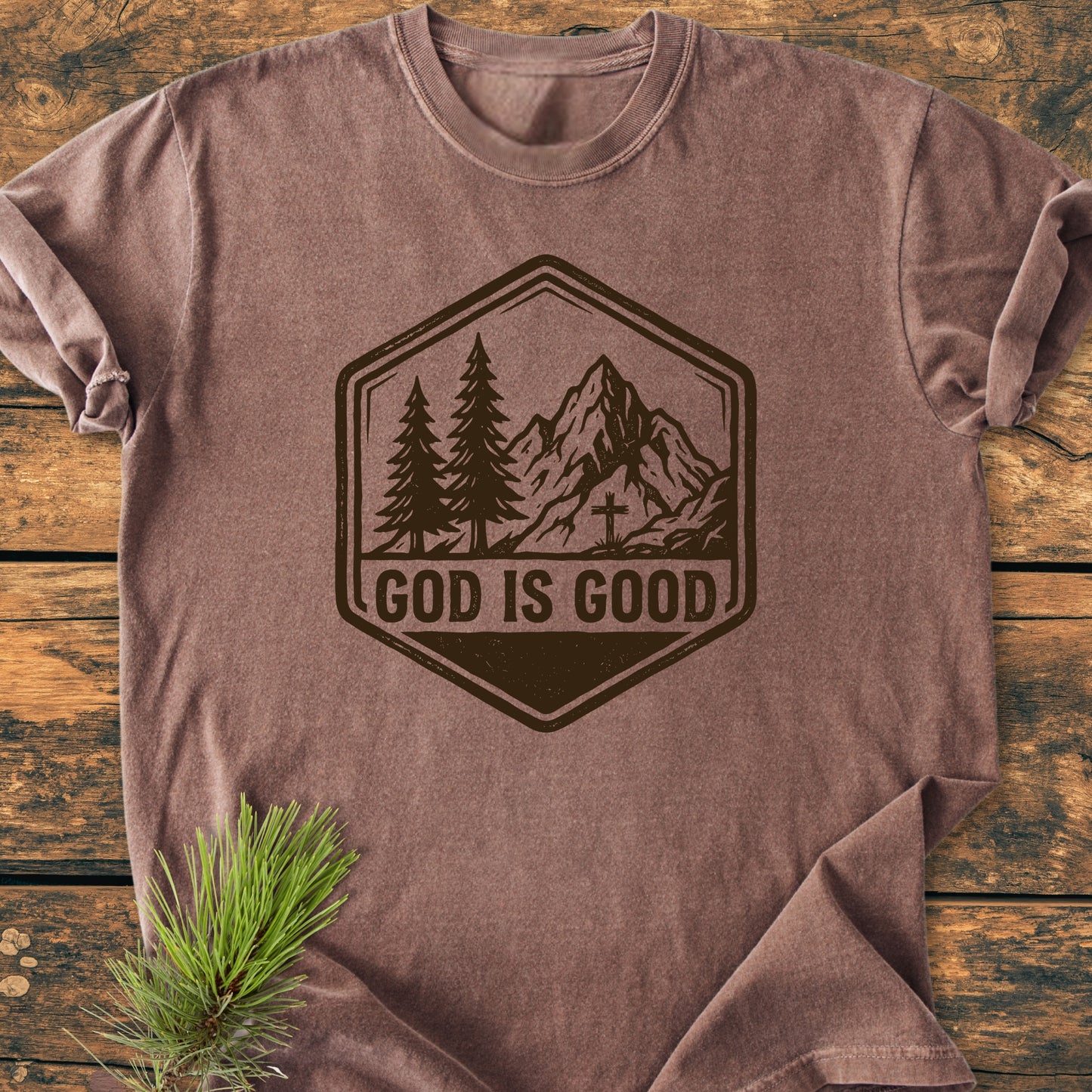 God Is Good - Vintage Wash Tee on wooden surface, featuring hexagonal badge with mountains and trees. GOD IS GOOD text below. Pine sprig accentuates this soft, durable, relaxed-fit garment-dyed shirt.