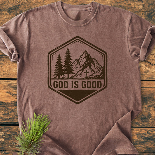 God Is Good - Vintage Wash Tee on wooden surface, featuring hexagonal badge with mountains and trees. GOD IS GOOD text below. Pine sprig accentuates this soft, durable, relaxed-fit garment-dyed shirt.