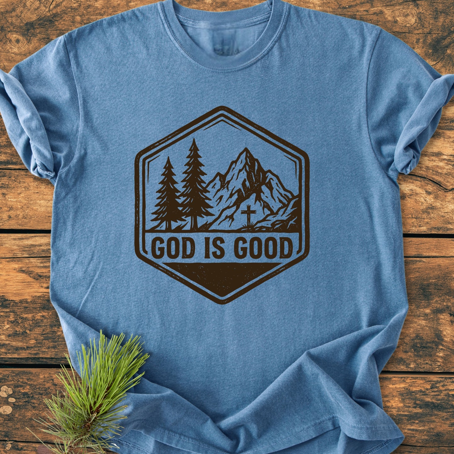 God Is Good - Vintage Wash Tee featuring a rustic badge graphic with pine trees and a mountain peak, displayed on a wooden surface with a pine sprig beside it.