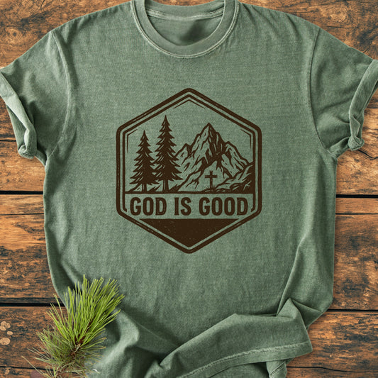 God Is Good - Vintage Wash Tee: A heather-green T-shirt with a badge-style graphic of trees and a mountain, laid on wood, featuring “GOD IS GOOD” text, highlighting comfort and durability.