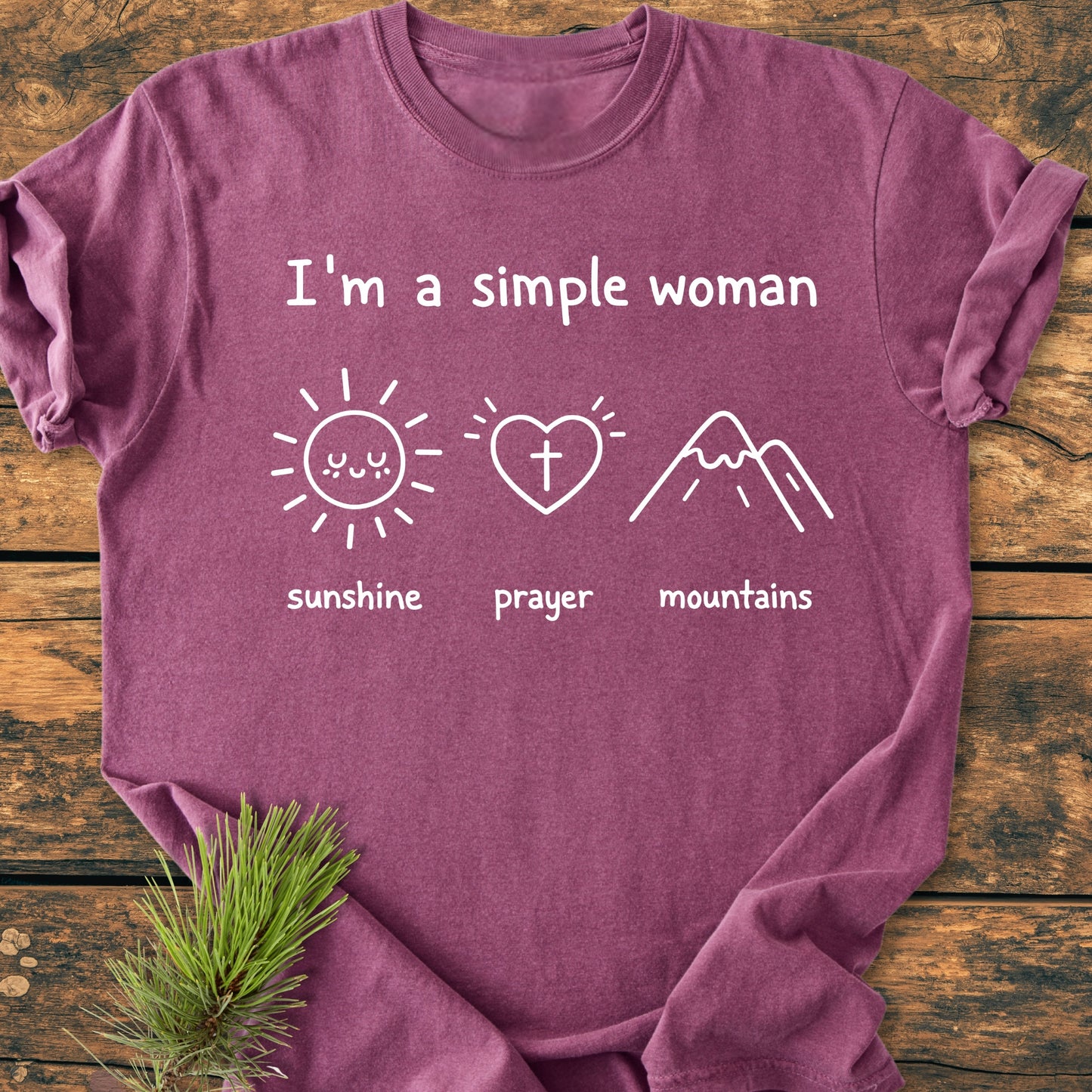 I'm A Simple Woman - Sunshine Prayer Mountains - Vintage Wash Tee, featuring white print icons on plum-purple fabric, laid on a wooden surface with an evergreen sprig.