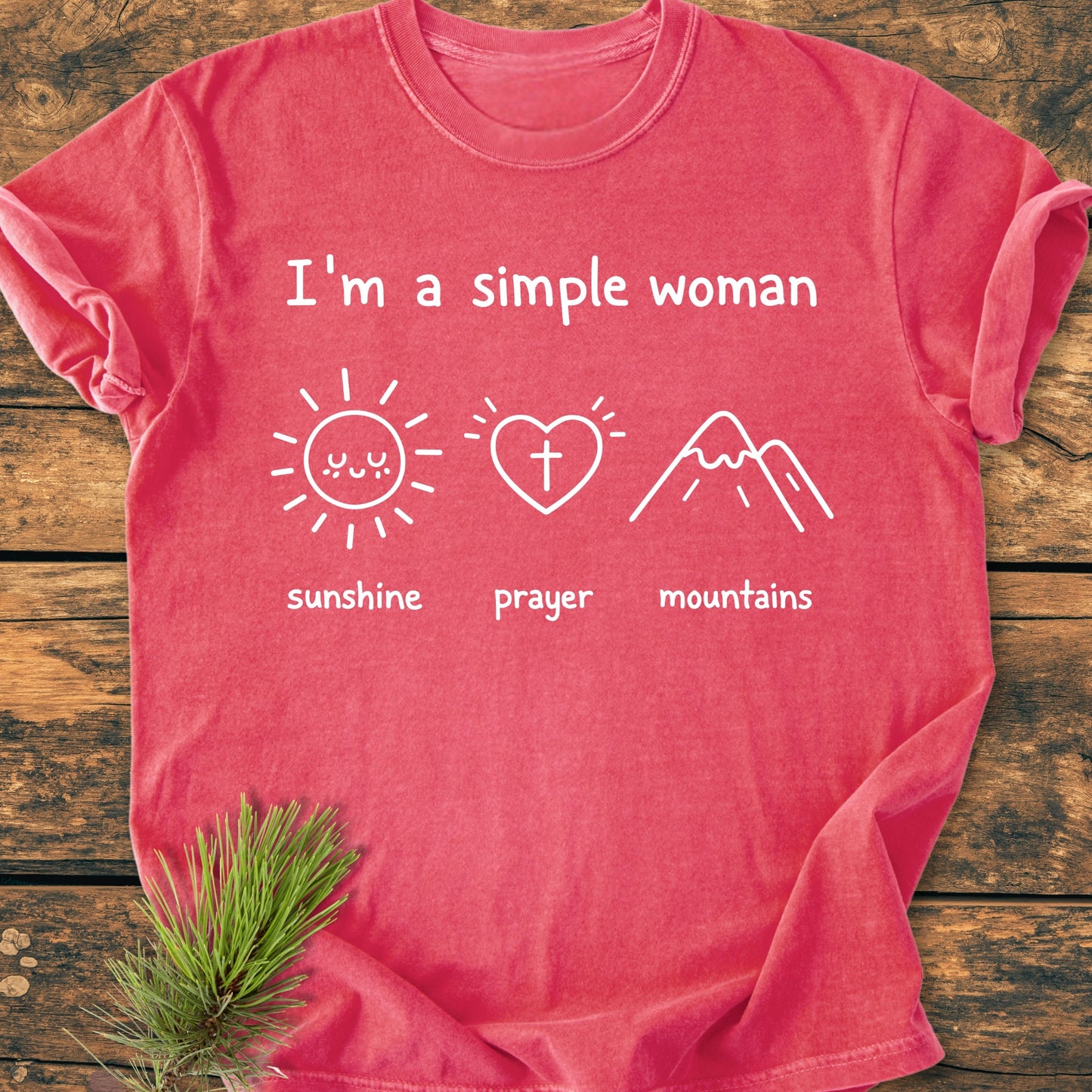I'm A Simple Woman - Sunshine Prayer Mountains - Vintage Wash Tee laid flat, featuring white text and icons on reddish-coral fabric. Displayed on a wooden surface with a pine sprig accent.