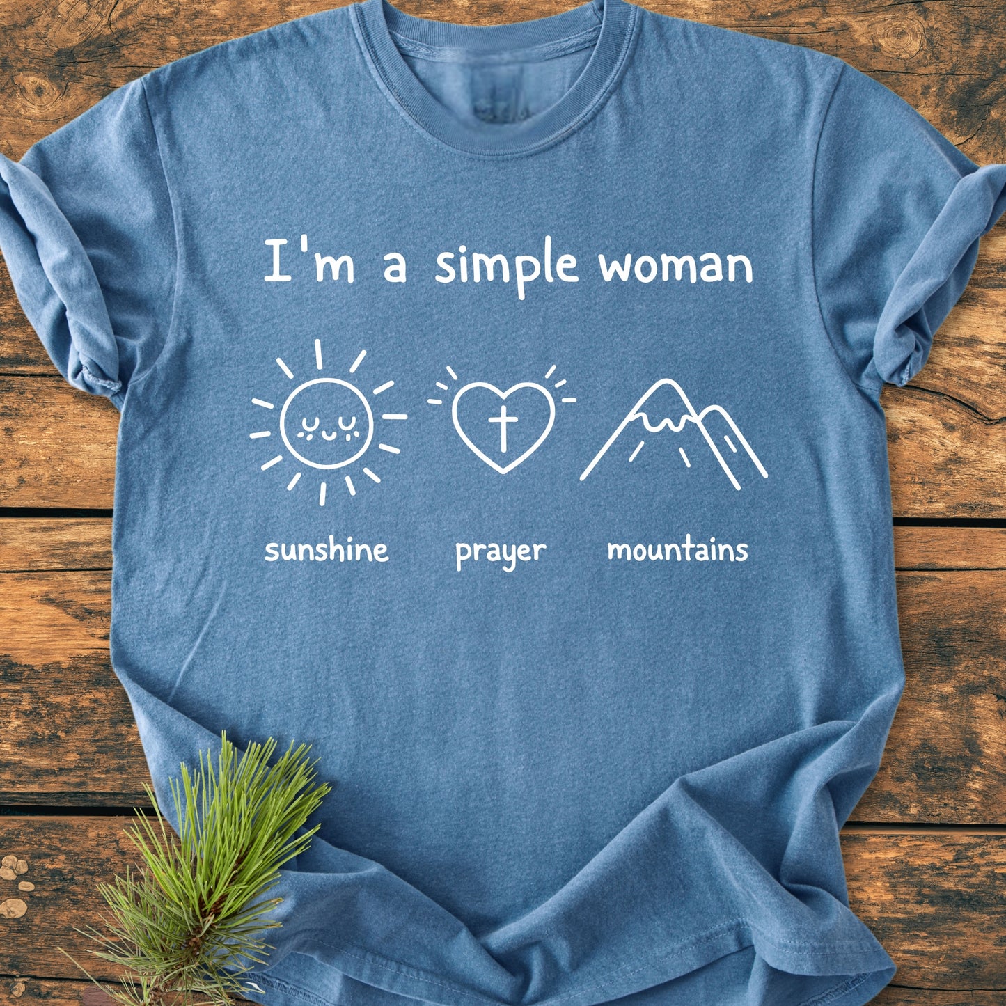 I'm A Simple Woman - Sunshine Prayer Mountains - Vintage Wash Tee features minimalist white line drawings of a sun, heart, and mountain on a heather-blue T-shirt laid on wood.