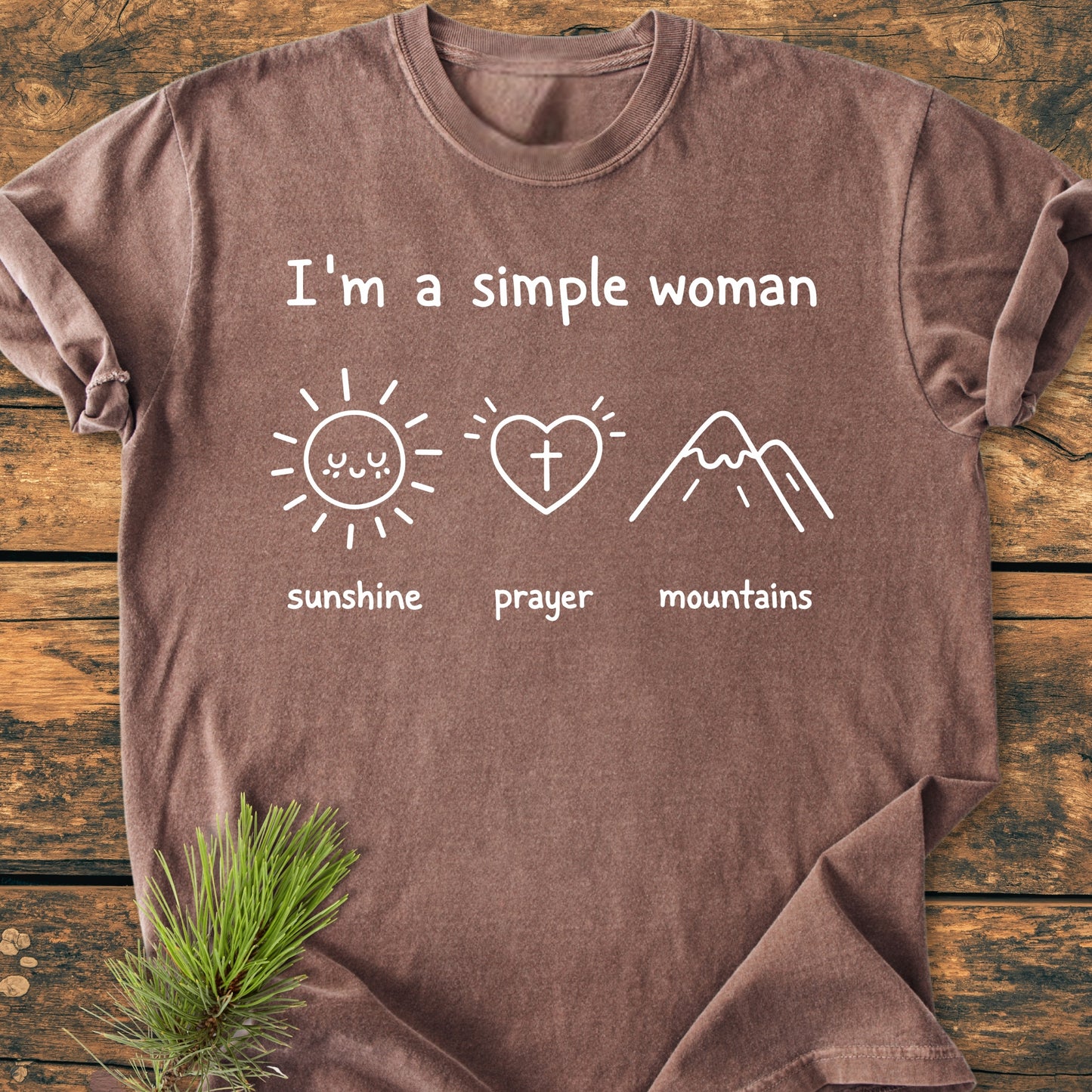 I'm A Simple Woman - Sunshine Prayer Mountains - Vintage Wash Tee, a brown T-shirt with white text and icons, laid flat on wood, featuring a pine sprig accentuating its rustic charm.