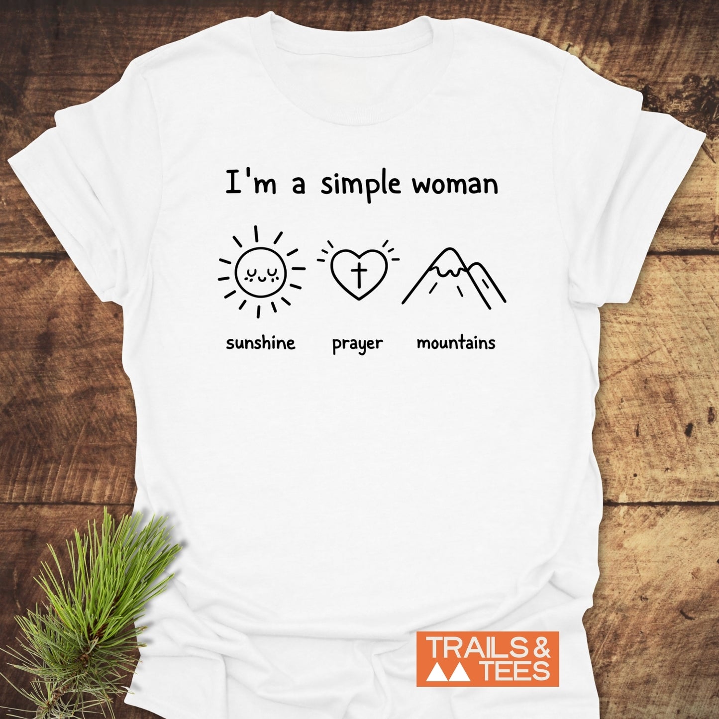 A Simple Woman - Sunshine Prayer Mountains T-Shirt displayed flat on wood, featuring icons of a sun, heart with cross, and mountains. Includes TRAILS & TEES. logo, highlighting outdoor adventure spirit.