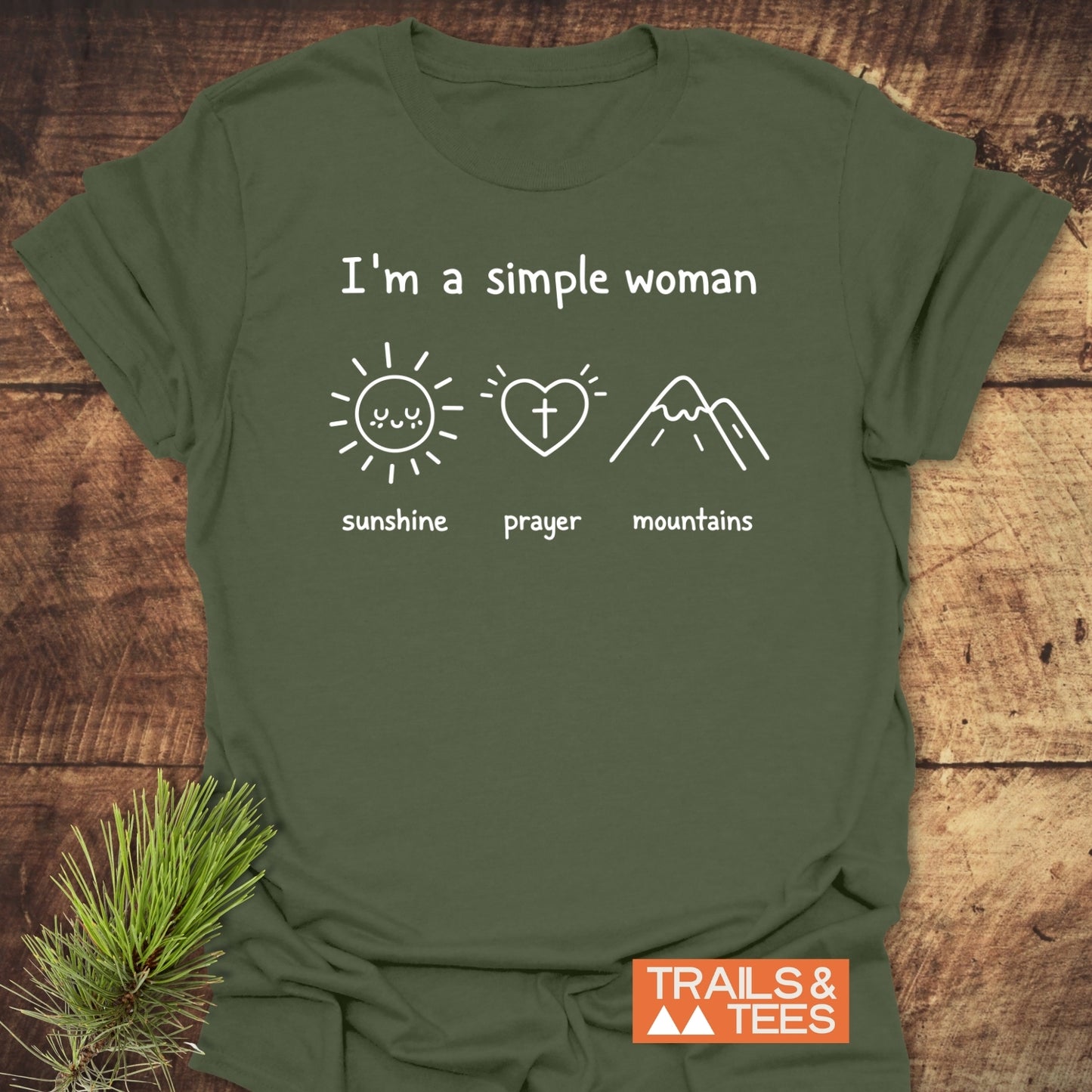A Simple Woman - Sunshine Prayer Mountains T-Shirt with icons of a sun, heart, and mountains on olive-green fabric, featuring a small TRAILS & TEES logo, laid on a wooden surface.