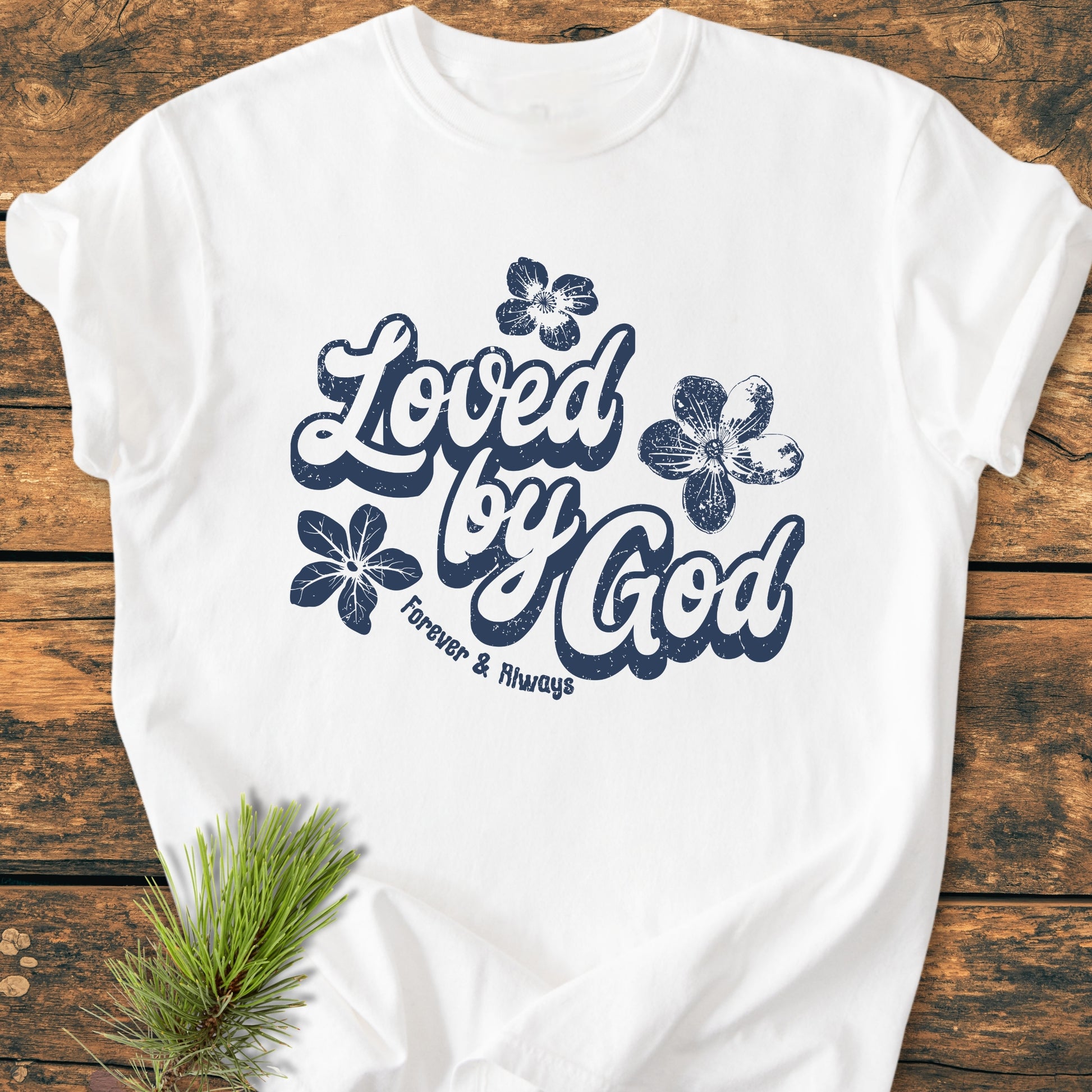 Loved By God - Vintage Wash Tee: A white tee with Loved by God in navy script, featuring distressed floral accents. Laid on rustic wood, showcasing its soft, relaxed fit and garment-dyed texture.
