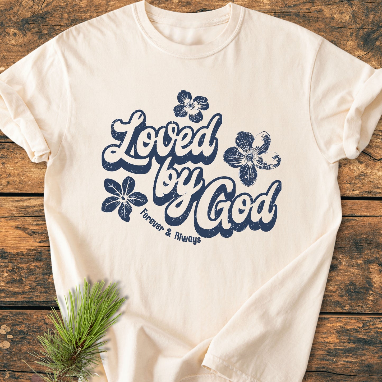 Loved By God - Vintage Wash Tee features a navy-blue script and floral design on a cream T-shirt, laid on wood, with a pine sprig accentuating its rustic appeal.