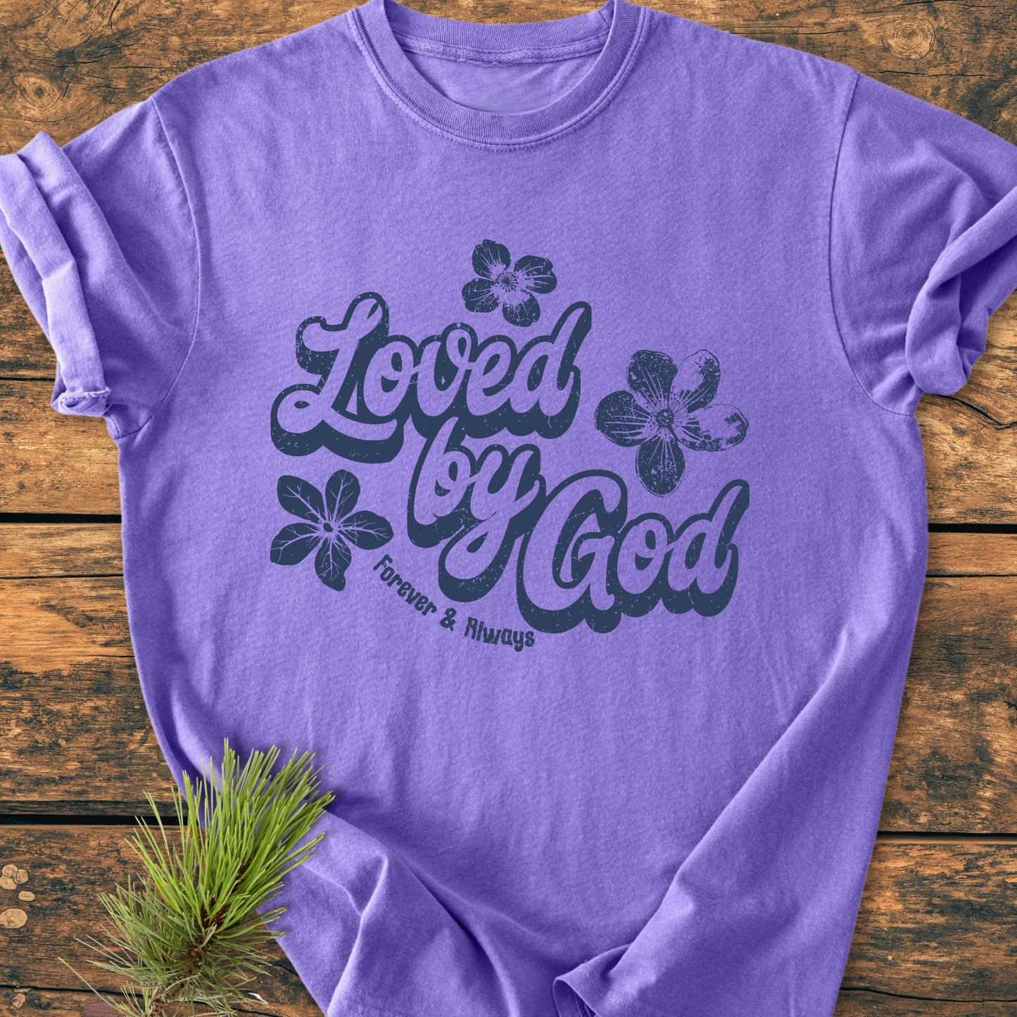 Loved By God - Vintage Wash Tee on wood, featuring bold script and flower accents, relaxed fit for comfort, made of soft, durable, pre-shrunk 100% ring-spun cotton, includes evergreen sprig detail.