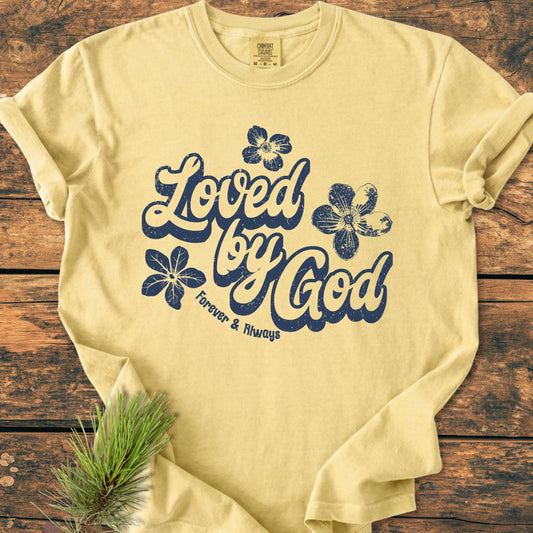 Loved By God - Vintage Wash Tee features a distressed navy script on cream fabric, adorned with floral motifs. Laid on wood, it includes a pine branch accent, showcasing its rustic charm.