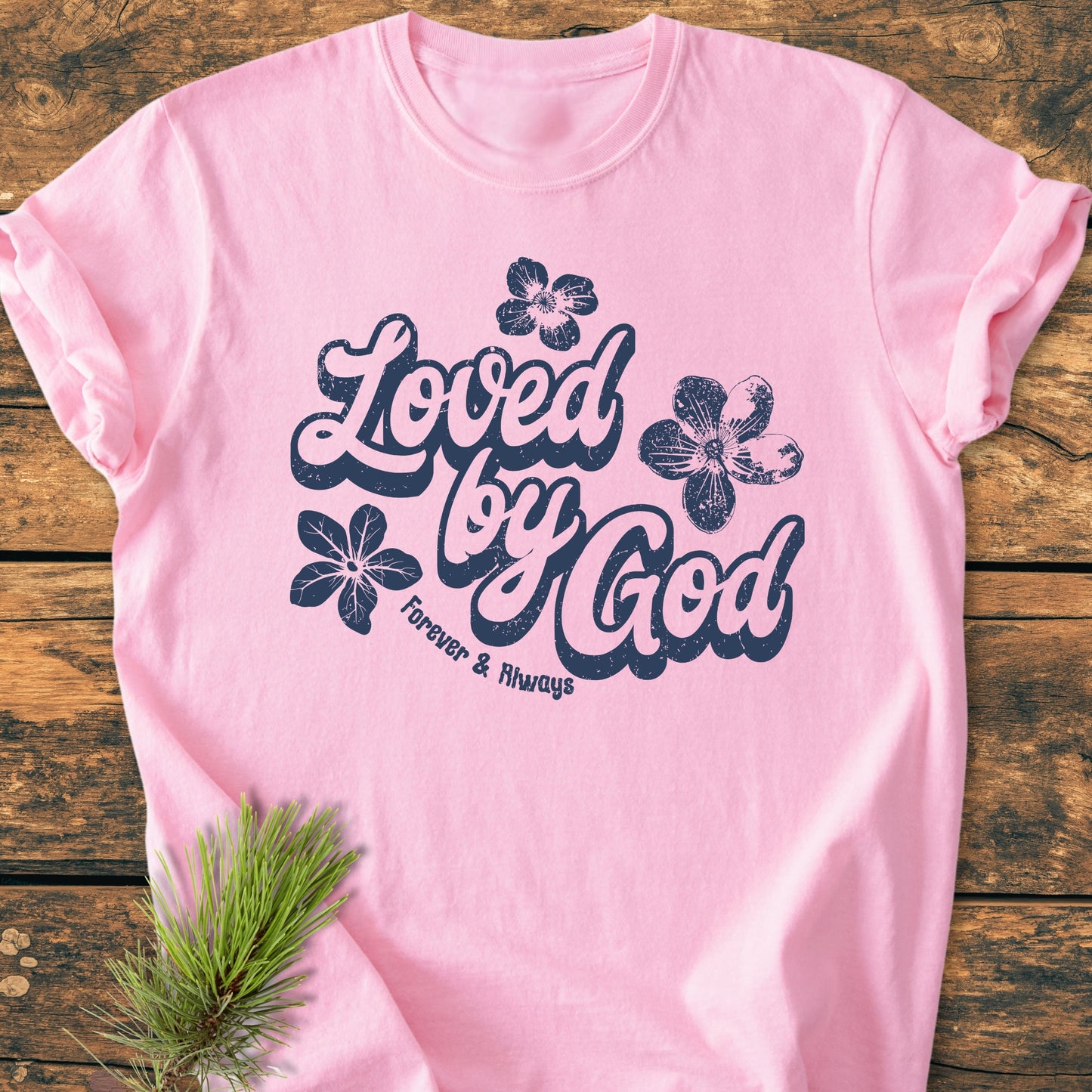 Loved By God - Vintage Wash Tee: A light-pink T-shirt with “Loved by God” and “Forever & Always” in script, adorned with flower illustrations, laid on a wooden surface with a pine sprig accent.