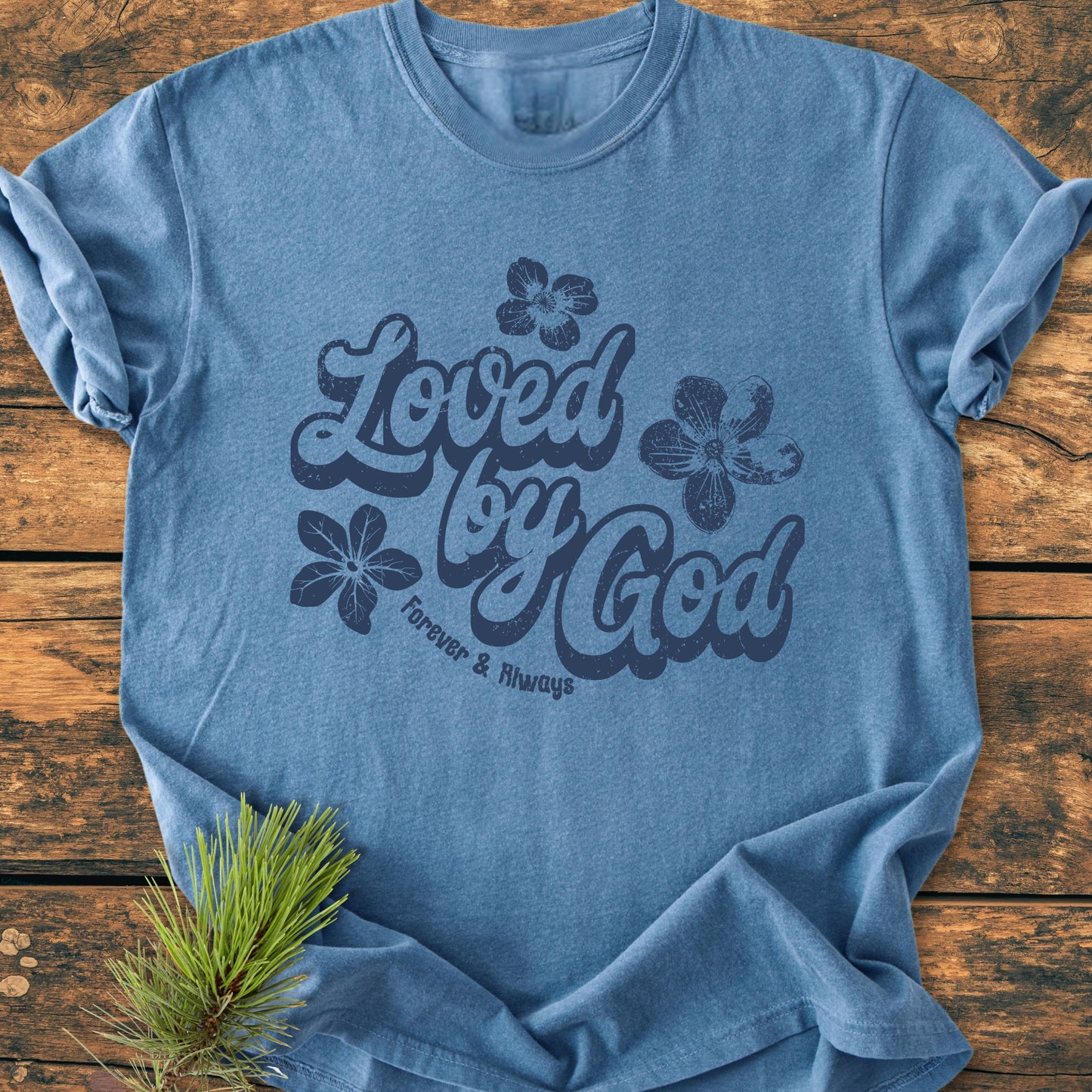 Heather-blue Loved By God - Vintage Wash Tee features dark-blue script and flower graphics, laid flat on wood. Garment-dyed cotton ensures comfort and durability, styled with a pine sprig for elegance.