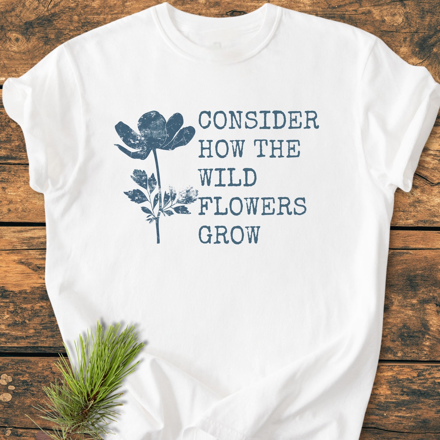 Consider How The Wildflowers Grow Luke 12,27 - Vintage Wash Tee: White T-shirt featuring a blue wildflower illustration and text, laid on wood with a pine sprig beside it, highlighting its relaxed, soft-washed design.
