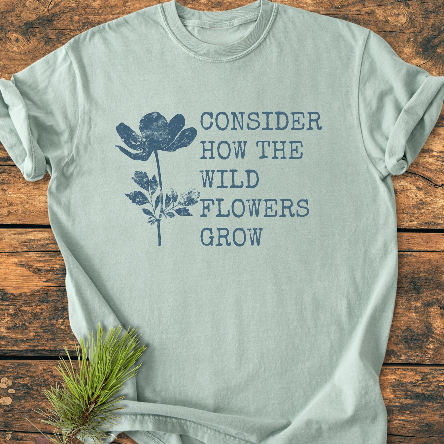 Consider How The Wildflowers Grow Luke 12,27 - Vintage Wash Tee in pale green, featuring a slate blue wildflower illustration and text. Laid flat on wood with pine needles accentuating its natural theme.