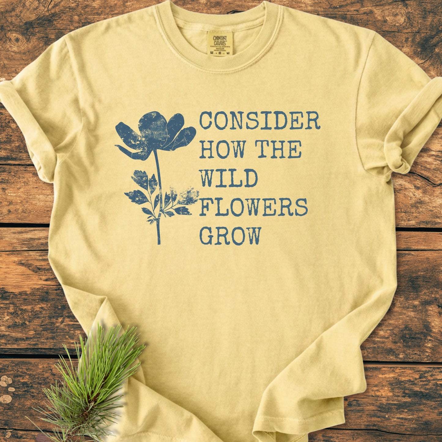 Consider How The Wildflowers Grow Luke 12,27 - Vintage Wash Tee in pale-yellow featuring a distressed wildflower graphic in slate-blue, laid out with a pine sprig on a wooden surface.