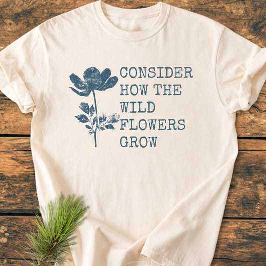 Cream-colored Consider How The Wildflowers Grow Luke 12,27 - Vintage Wash Tee, features a blue wildflower graphic. Laid on wood, styled with pine needles. Made from 100% ring-spun cotton for comfort.