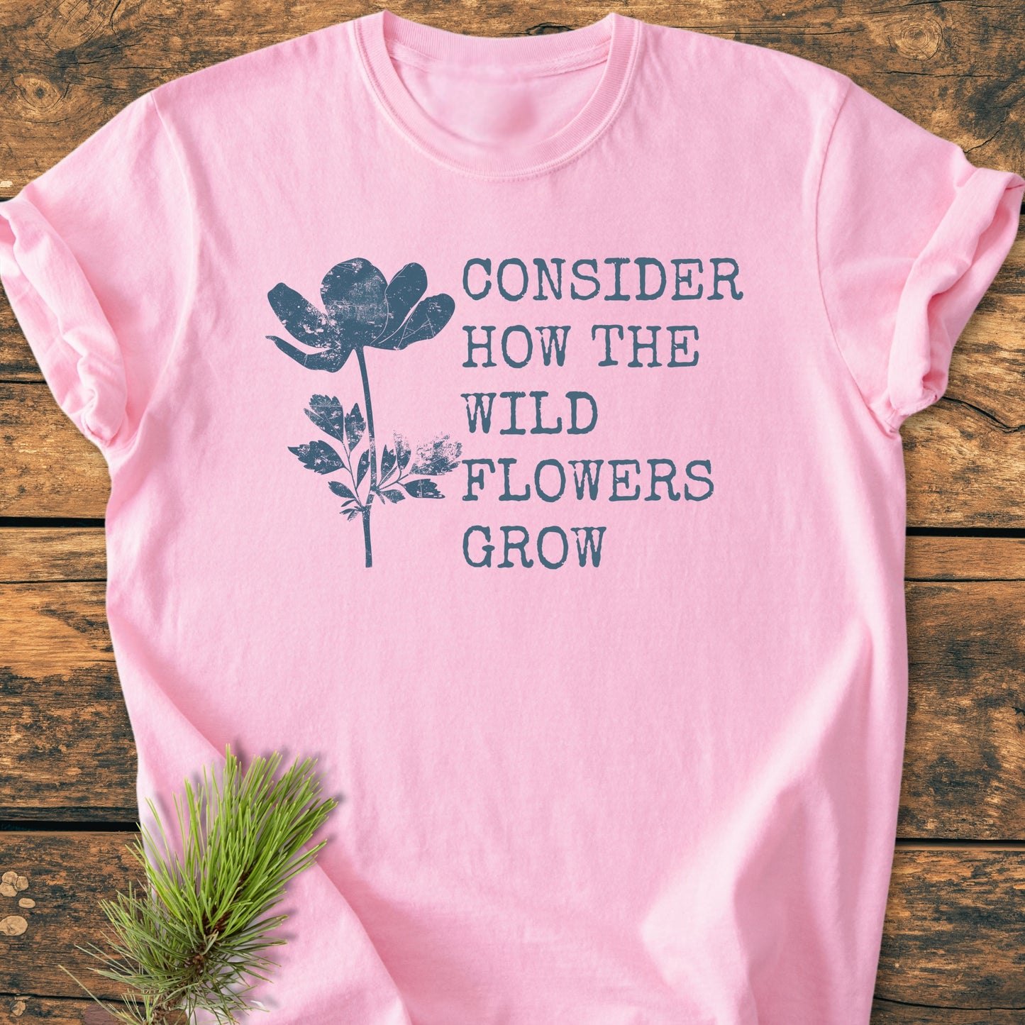 Pastel-pink Consider How The Wildflowers Grow - Vintage Wash Tee with distressed navy wildflower design, laid on wood. Features soft-washed cotton, relaxed fit, and decorative pine sprig.