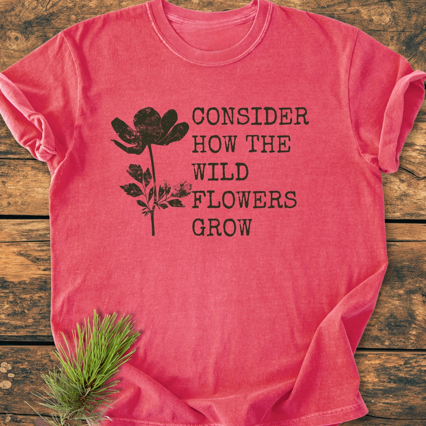 Consider How The Wildflowers Grow Luke 12,27 - Vintage Wash Tee features a black wildflower silhouette and quote on a pink/red shirt, styled with a pine branch for a rustic touch.