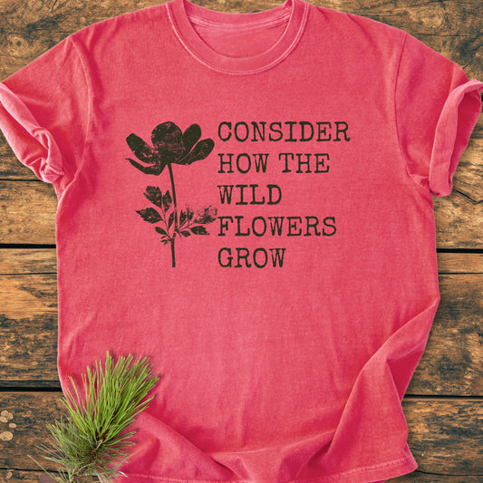 Consider How The Wildflowers Grow Luke 12,27 - Vintage Wash Tee features a black wildflower silhouette and quote on a pink/red shirt, styled with a pine branch for a rustic touch.