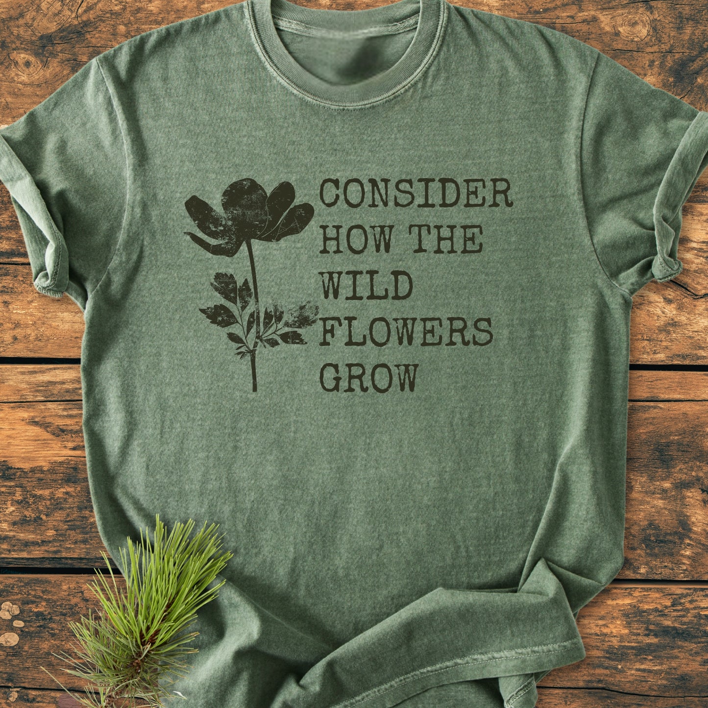 Consider How The Wildflowers Grow Luke 12,27 - Vintage Wash Tee in sage green features a black wildflower silhouette and quote, laid on wood. Includes pine sprig accent, enhancing its natural appeal.