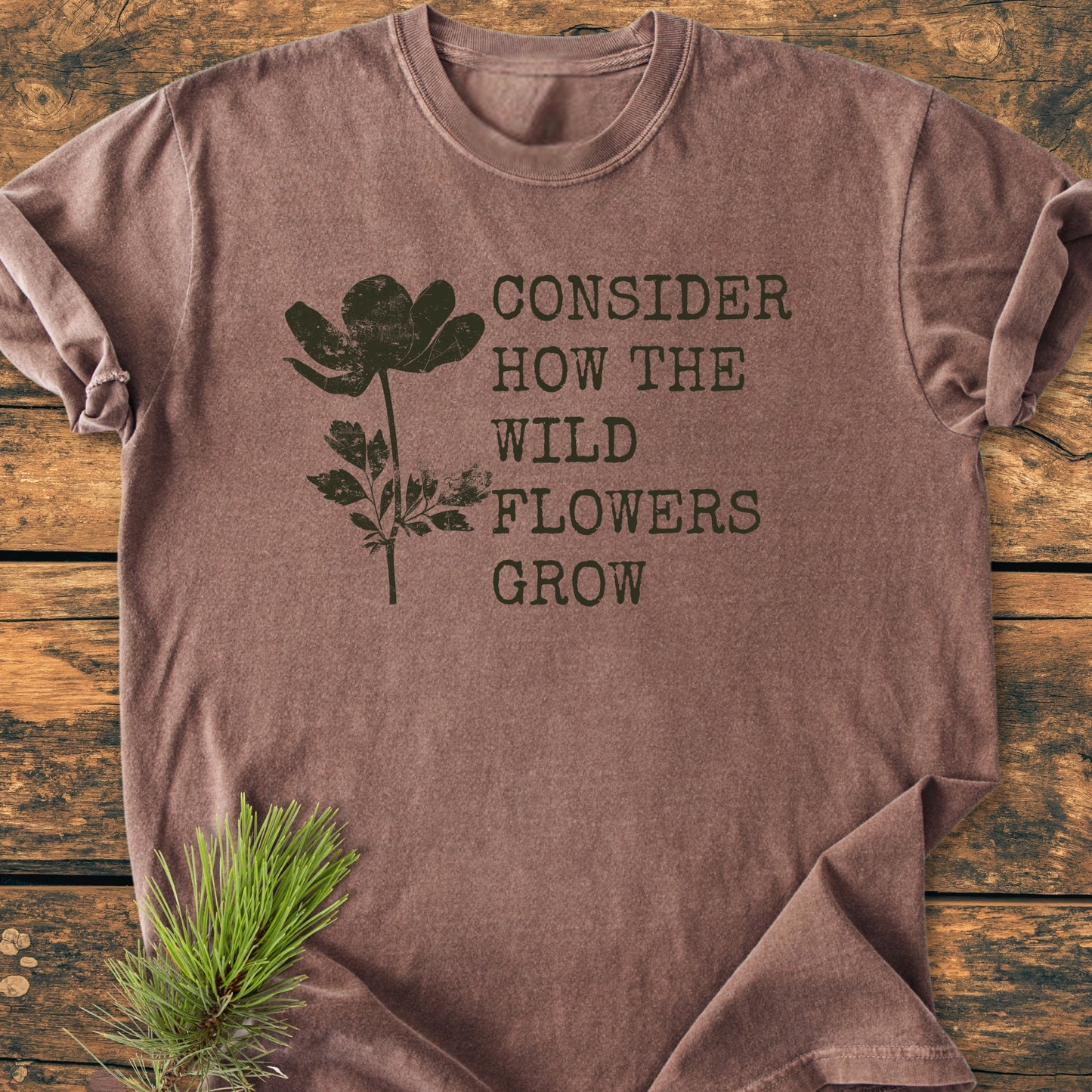Consider How The Wildflowers Grow Luke 12,27 - Vintage Wash Tee features a distressed wildflower graphic and text on a brown heathered T-shirt, laid flat with a pine sprig accent.